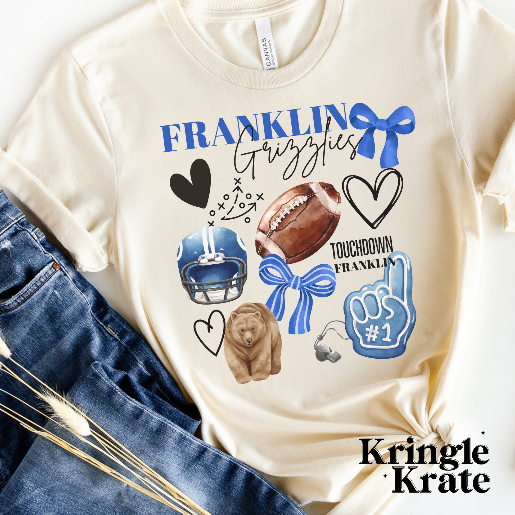 A cream-colored short-sleeve t-shirt featuring a vibrant, collage-style design that celebrates Franklin Grizzlies. The design includes football-themed elements such as a football, helmet, a play diagram, and a foam finger labeled "#1." Other fun graphics like a grizzly bear, a whistle, black hearts, and a blue bow are also displayed. The words "Franklin Grizzlies" are prominently printed in a mix of bold and cursive fonts, with "Touchdown Franklin" appearing alongside. The shirt is laid flat next to a pair 