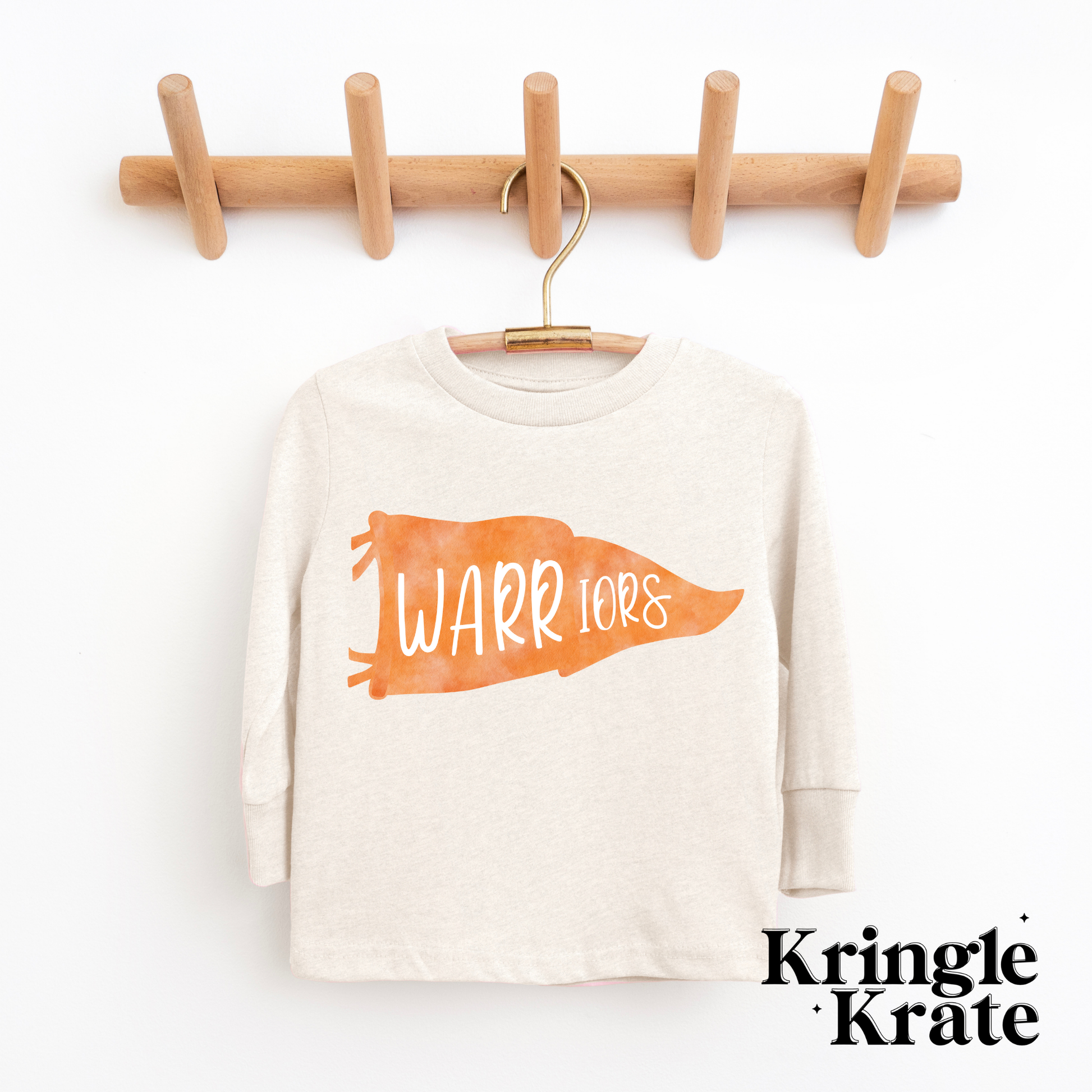 A natural heather-colored long-sleeve t-shirt featuring a bold orange pennant flag graphic with the words "Warriors" in playful white lettering. The shirt is displayed hanging on a wooden hanger against a white background, showcasing its simple yet spirited design. The t-shirt is a youth size, perfect for young fans, and has a casual, comfortable style ideal for showing school pride.